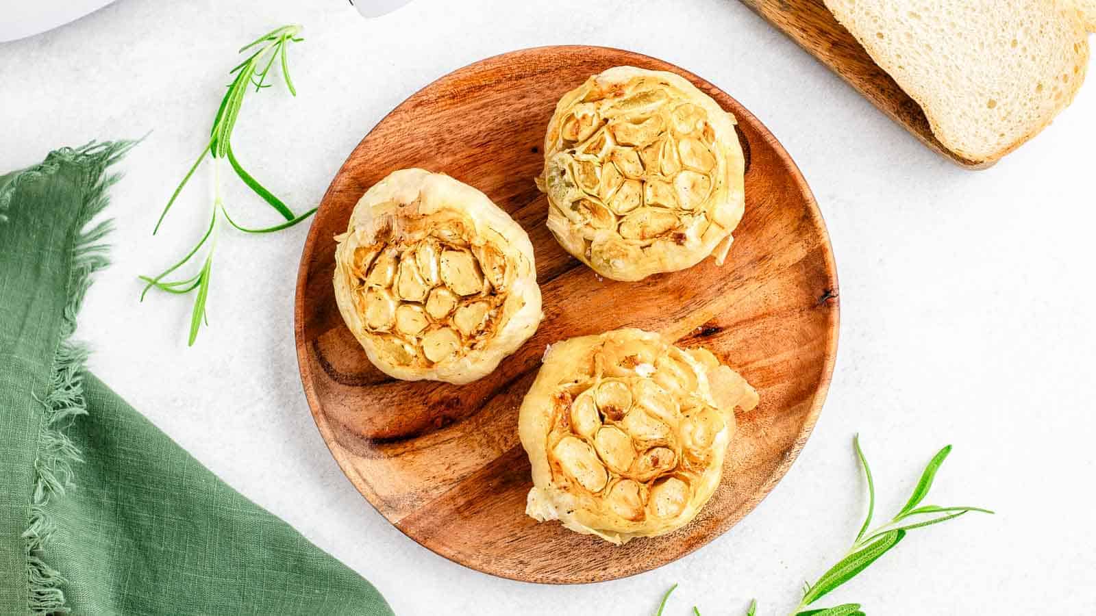 Air fryer roasted garlic on a wooden plate with bread and sprigs of rosemary.