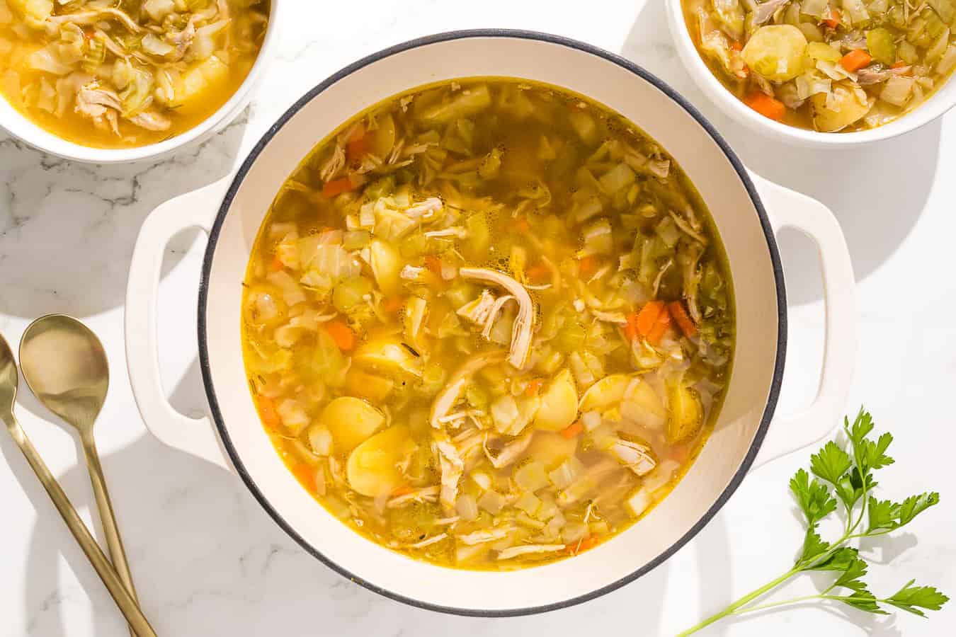 A bowl of chicken and cabbage soup is shown on a rustic wooden cutting board with parsley and a red linen.