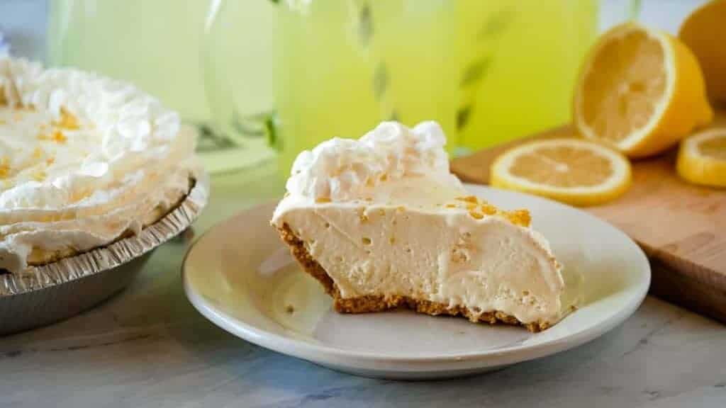 Slice of lemonade pie on a plate with lemonade in the background.