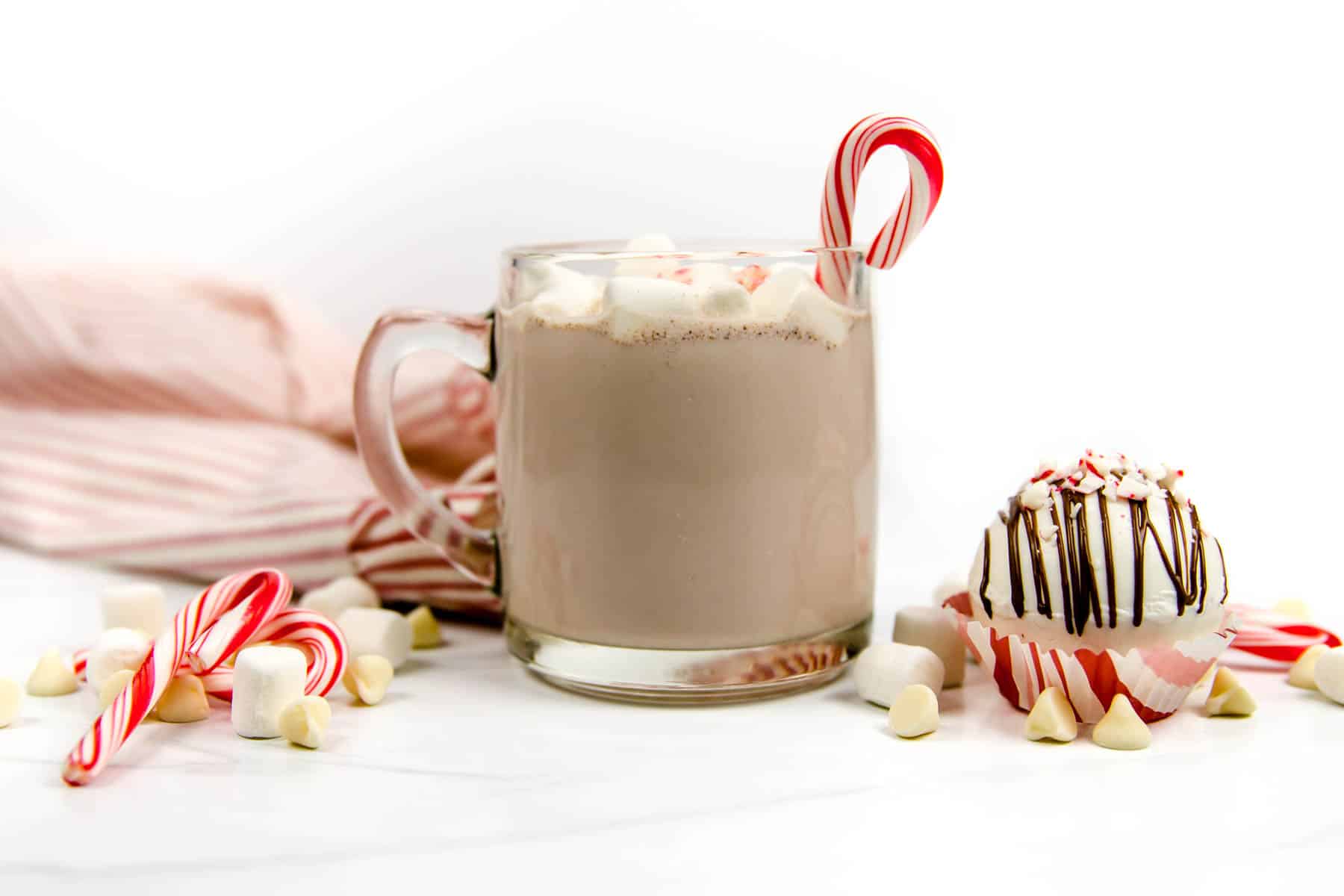 Image shows Ingredients for a peppermint hot cocoa bomb and a finished bomb sit around a mug of cocoa garnished with marshmallows and a candy cane.