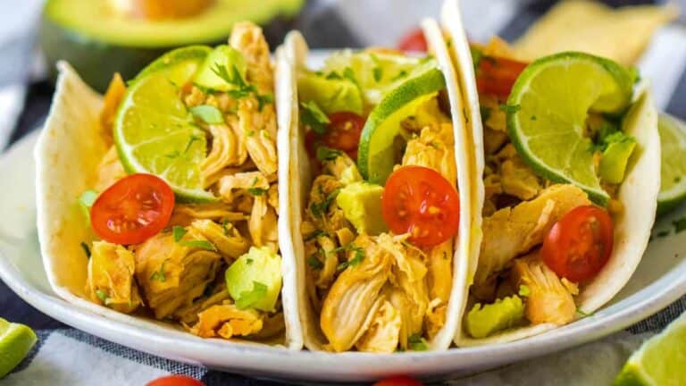 Three chicken tacos on a plate with tomatoes and avocado.