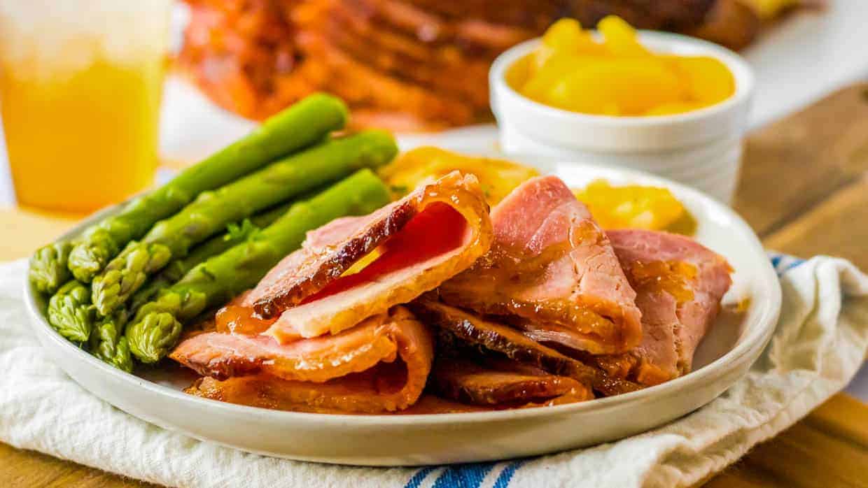 Spiral ham on a plate with asparagus and peaches in the background.