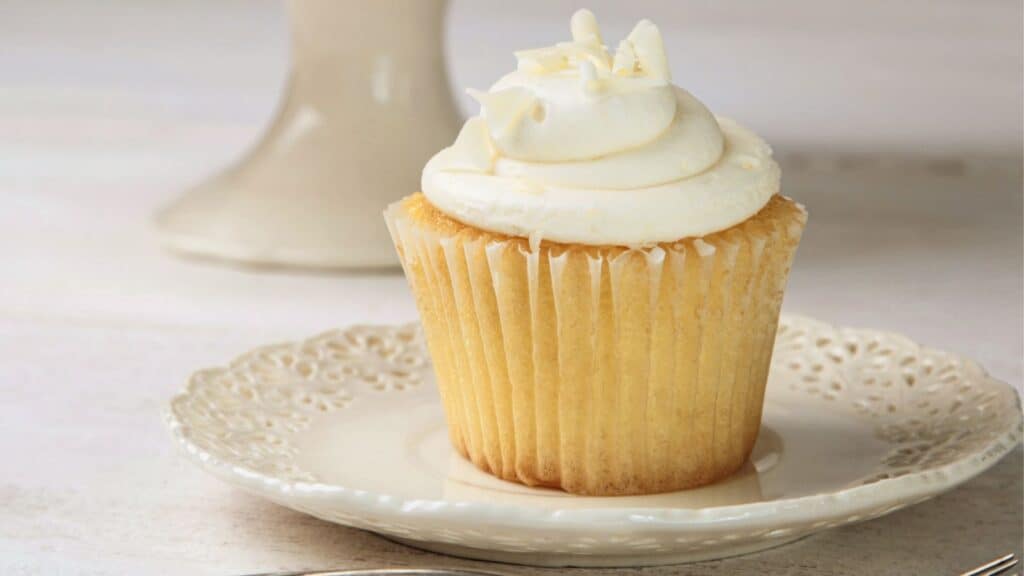 A white cupcake sits on a plate next to a fork.