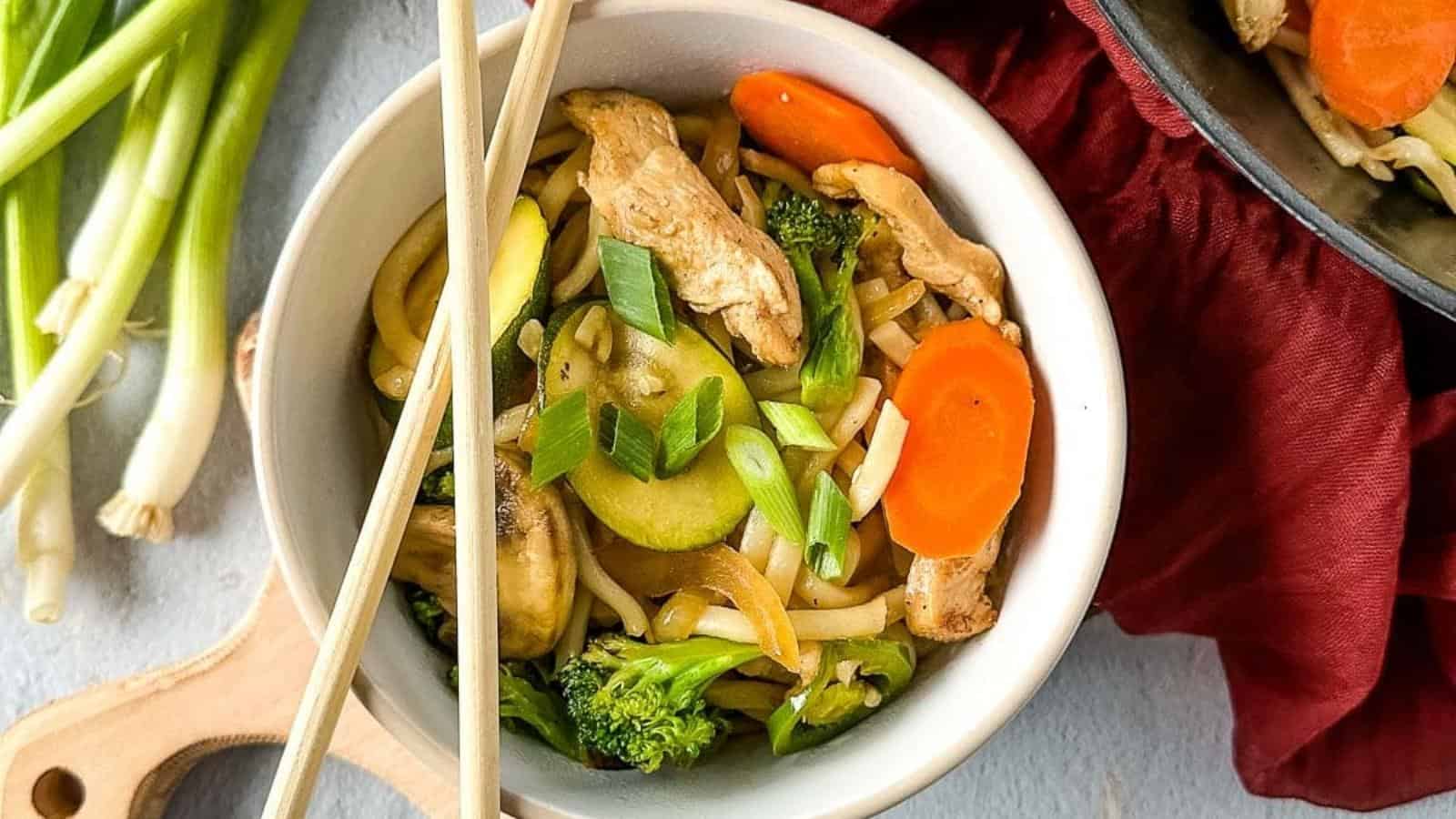 chicken yaki udon in a white bowl with chopsticks beside a wok.