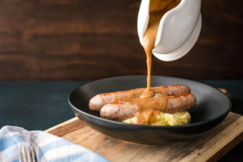 Gravy being poured over a plate of sausages and mashed potatoes.