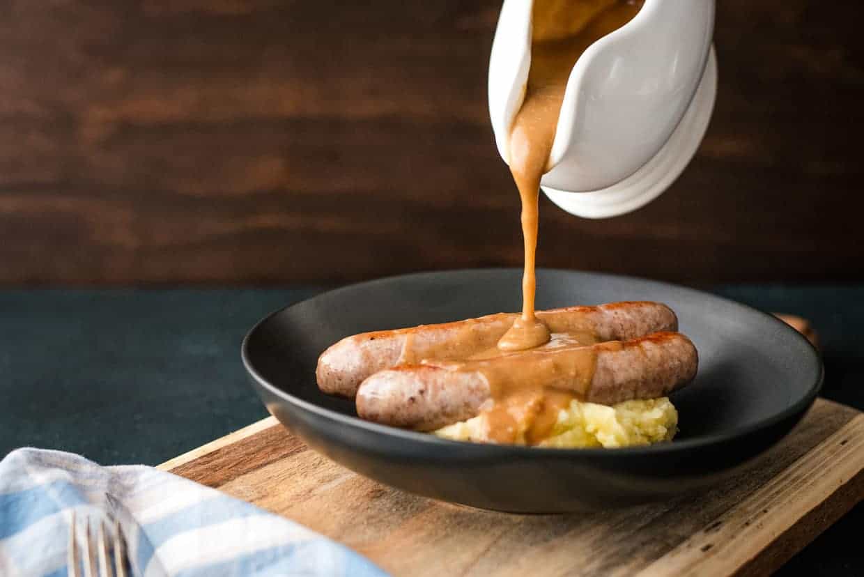 Gravy being poured over a plate of sausages and mashed potatoes.