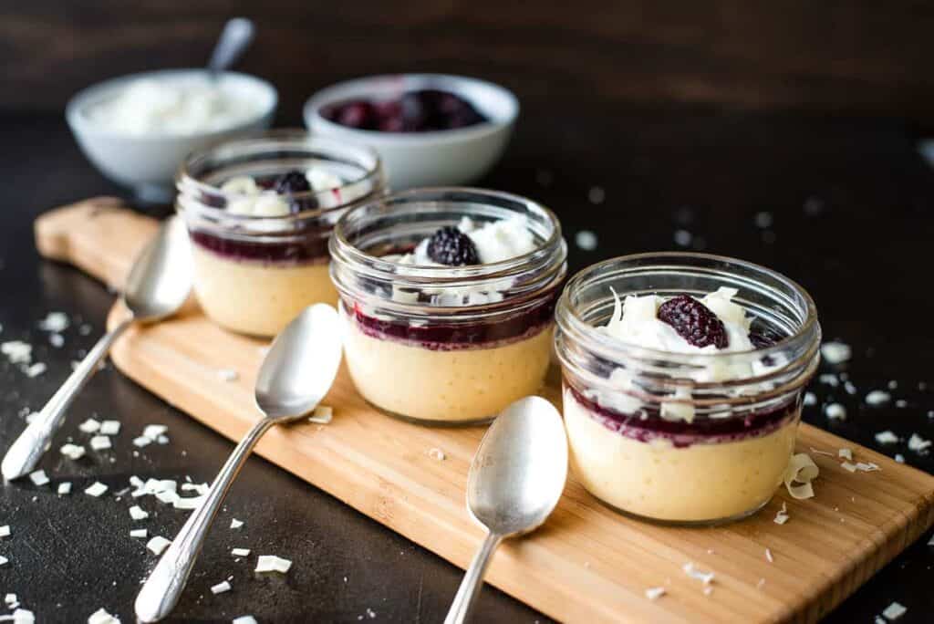 Three jars of blackberry cheesecake with spoons on a wooden cutting board, accompanied by decadent chocolate treats.