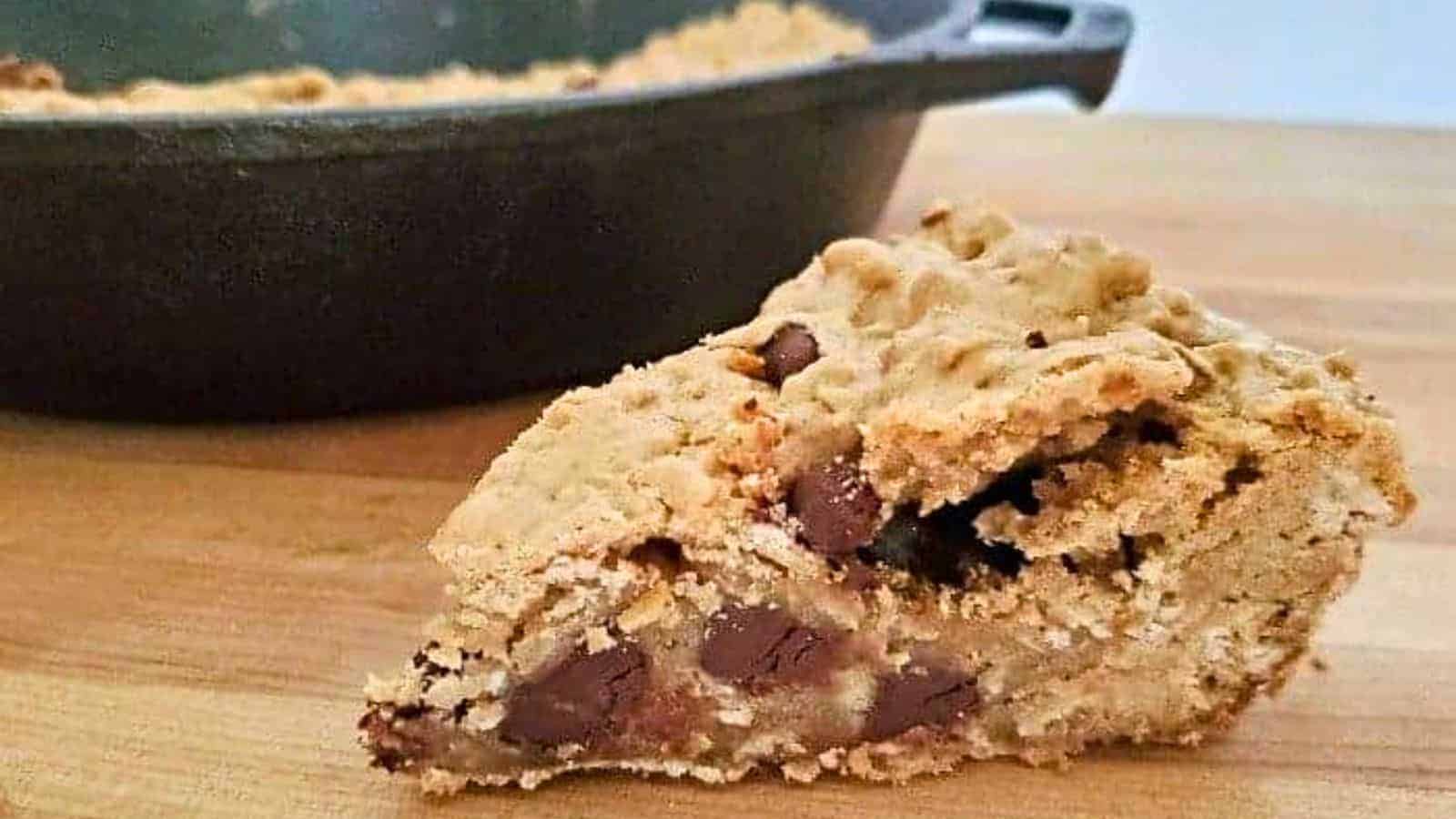 Image shows A piece of chocolate chip skillet cookie in front of a cast iron pan.