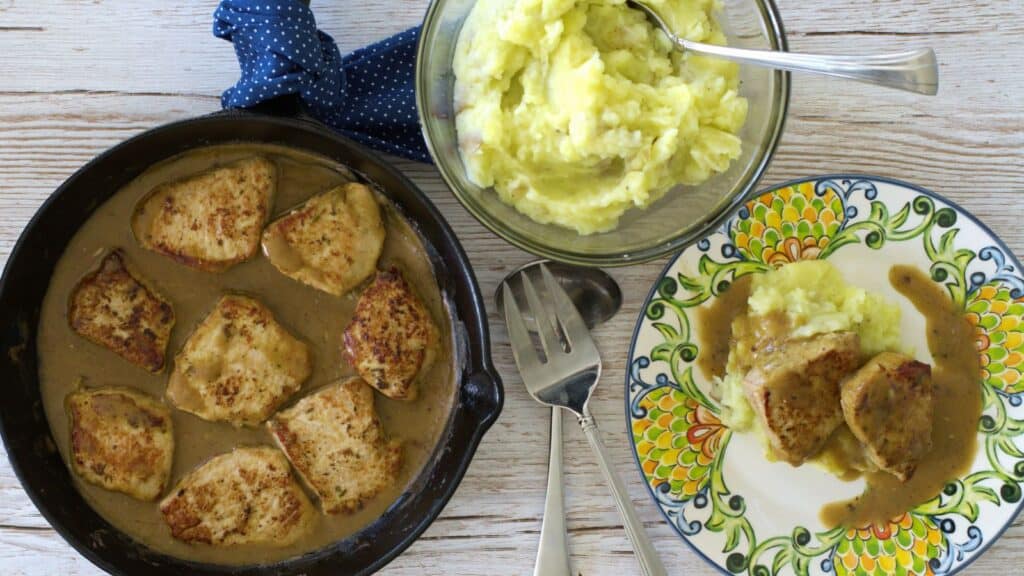 A plate of pork cutlets with gravy and mashed potatoes next to serving dishes.