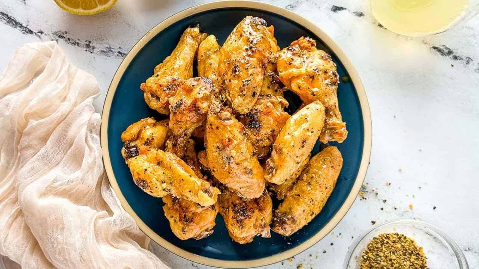 Hot honey lemon pepper wings on a dark blue plate surrounded by lemon pepper, lemons, a white linen, and lemonade.