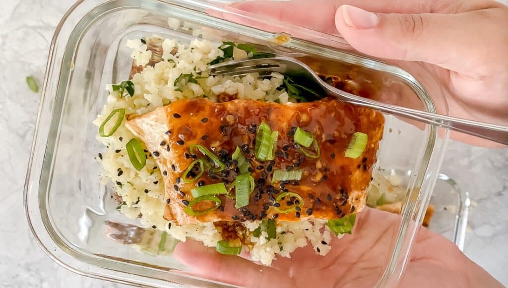 A person holding a glass container with cauliflower rice and salmon.