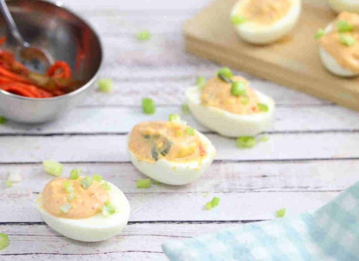 Kimchi spicy deviled eggs lined up with green onions on top and a bowl of kimchi behind them with more eggs on a cutting board.