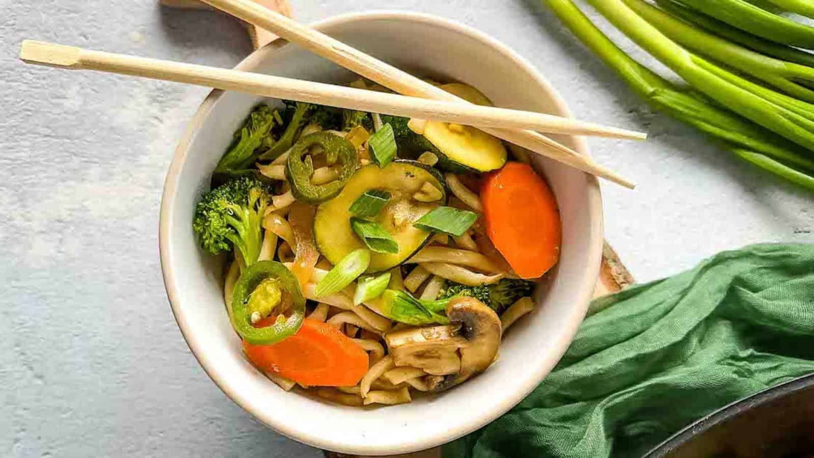 A bowl of vegetable yaki udon is shown with wooden chopsticks, a wok of yaki udon, and scallions in the background.