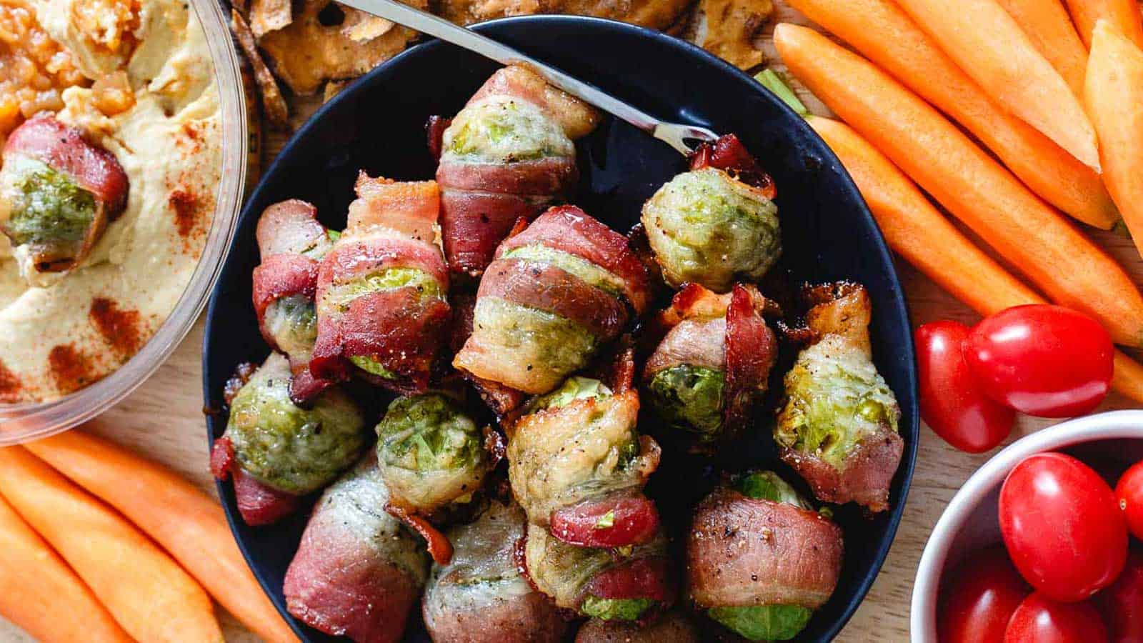 Bacon wrapped Brussels sprouts on a dark plate with vegetables surrounding it.