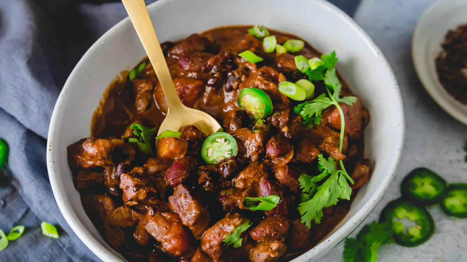 A bowl of lamb chili  garnished with cilantro, jalapeños and green onions with a gold spoon.