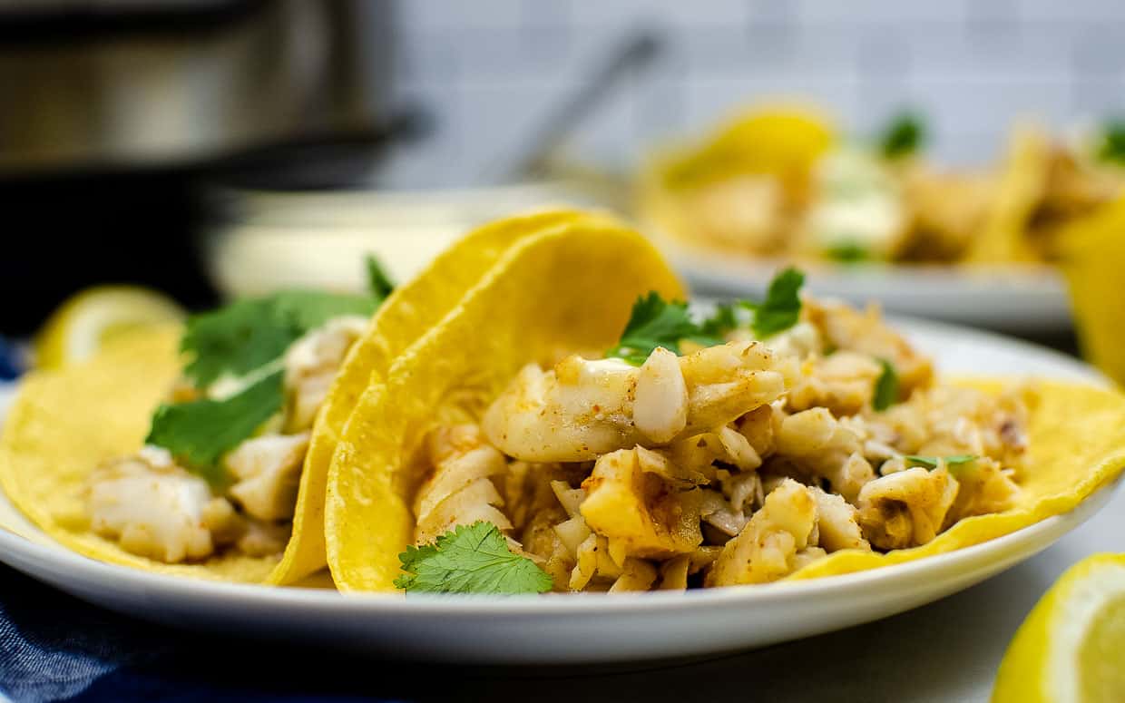 Fish tacos on a plate in tortillas with coriander and lemon wedges.