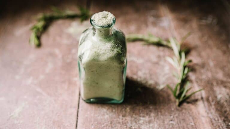 A bottle of green powder with rosemary sprigs on a wooden table.